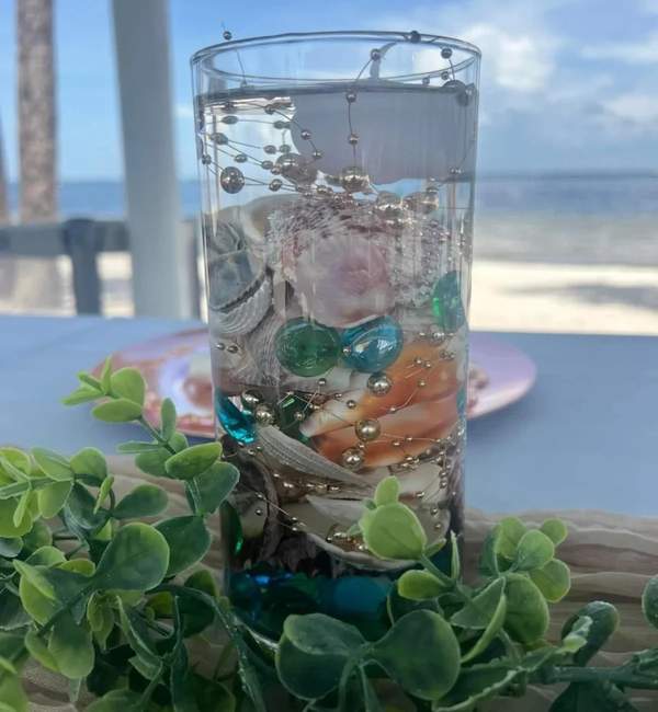 A decorative glass cylinder centerpiece filled with sea shells and gold beads on a table overlooking the beach