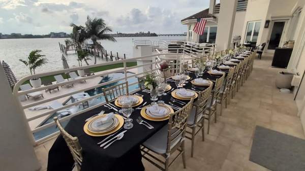 A long banquet dining table set with gold charger plates and black tablecloths on a waterfront balcony overlooking a canal